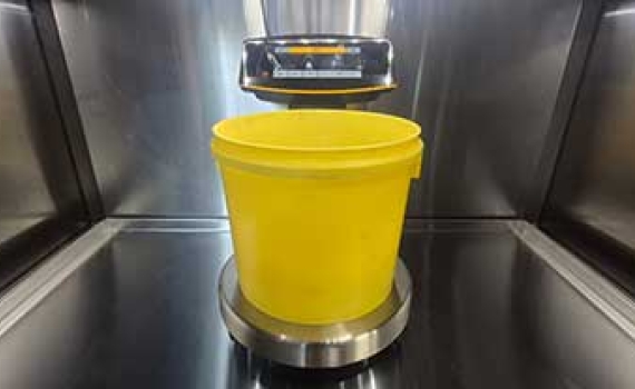 A yellow bucket sits on a round metal scale inside a stainless steel chamber, with equipment mounted above.