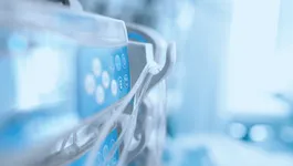 Close-up of a medical IV infusion pump with circular buttons and attached tubing, set against a blurred clinical background with cool blue lighting.