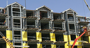 A multi-story building under construction with an exposed metal frame. The lower floors have bright yellow insulation panels, and an orange construction lift is in front.