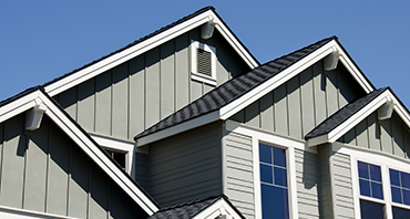 Close-up of a house roofline with gray siding, dark shingled gabled roof, and white trim, set against a clear blue sky.