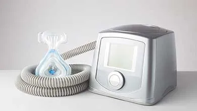 Silver CPAP machine with digital screen, gray coiled tube, and clear nasal mask.