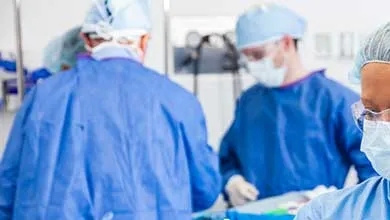 Surgeons in blue gowns, masks, and caps operate in a sterile room, a gloved hand prominent in the foreground.