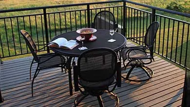 A round black patio table with four matching chairs sits on a wooden deck with a black railing, overlooking a grassy field at sunset.