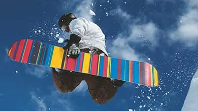 A snowboarder airborne above a snowy slope, with a colorful snowboard against a bright blue sky.