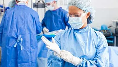 A female surgeon in a blue gown, mask, and cap puts on sterile gloves in an operating room, with other surgeons working in the background.