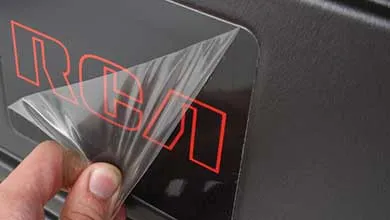 一只手正在从黑色塑料表面揭下一层透明保护膜，下面有红色字母。