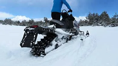 A person in a blue jacket rides a snowmobile across snowy ground, with trees and distant mountains in the background.