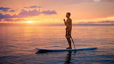 A man stands on a paddleboard on calm water at sunset.