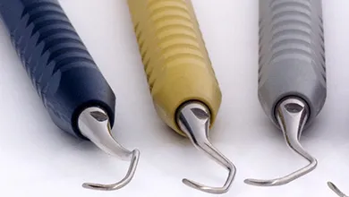 Three dental instruments with ribbed handles in dark blue, yellow, and gray, each with a curved metal hook tip, laid horizontally on a white background.