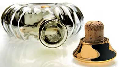 A clear, fluted glass bottle lies on its side beside its stopper, which has a cork and a gold, bell-shaped base with a black wavy pattern.