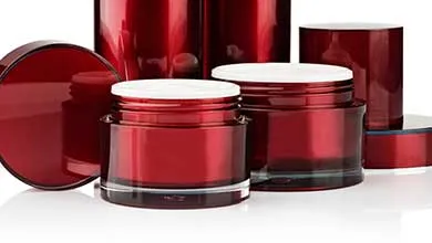 Glossy red cosmetic containers on a white background, with two open round jars showing white interiors in front, and taller cylindrical bottles behind. One jar lid rests on its side.