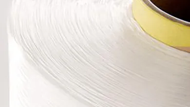 Close-up of a large spool of white thread tightly wound around a tan cardboard core, showing the smooth, linear texture of the strands.