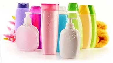 Various colorful personal care bottles, including two pump dispensers—one peach and one white—in front of taller cylindrical bottles in purple, pink, blue, green, and yellow. Pink flowers are on the left and a rolled yellow towel is on the right, all set against a white background.