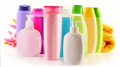 Colorful personal care bottles of various shapes and sizes, including pump dispensers and tall cylinders in purple, pink, blue, green, and yellow, arranged with a folded yellow towel and pink flower on a white background.