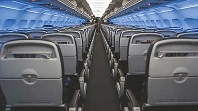 Empty commercial airplane cabin interior with rows of gray seats along a central aisle, overhead storage bins, and blue ambient lighting.