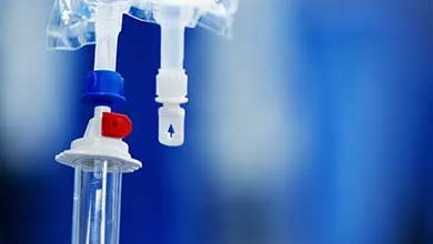 Close-up of IV infusion equipment showing a clear drip chamber, red roller clamp, blue connector, and white injection port with arrow, hanging from a transparent IV bag against a blurred blue background.