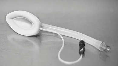 White medical tracheostomy mask with an oval cushioned facepiece, clear flexible tubing, and a small black clamp connector, resting on a smooth gray metallic surface.