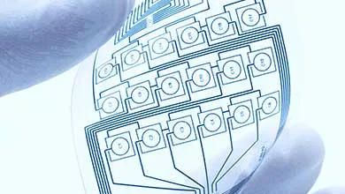 A flexible transparent sheet with circular electronic sensors and circuit traces, held between gloved fingers.