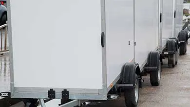 A row of white enclosed cargo trailers parked side by side on concrete, showing smooth exterior panels, black door handles, wheels, and rear hitches.