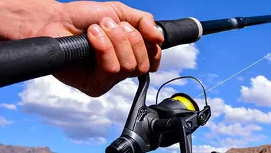 Close-up of a hand holding a black fishing rod with a spinning reel against a blue sky with white clouds.