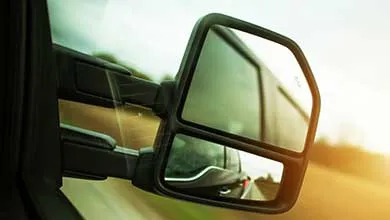 Close-up of a black vehicle side mirror with two sections: a main rectangular mirror and a smaller convex spotter mirror below, both reflecting the surroundings. The background is golden-lit and motion-blurred, indicating the vehicle is moving.